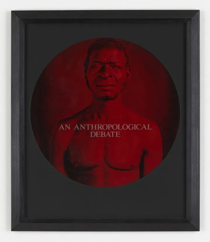 Fotografia storica virata in rosso, entro una cornice circolare su fondo nero: un uomo nero a torso nudo ripreso frontalmente, con una scritta sovrimpressa al centro.
